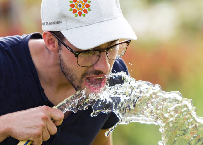 An image of a young man drinking from a water hose with an Outside Garden hat on