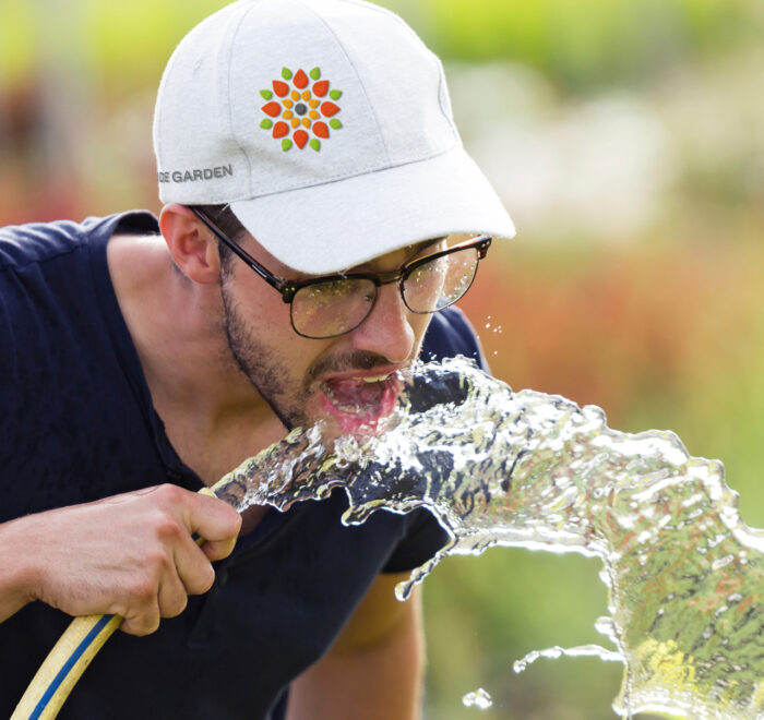 An image of a young man drinking from a water hose with an Outside Garden hat on
